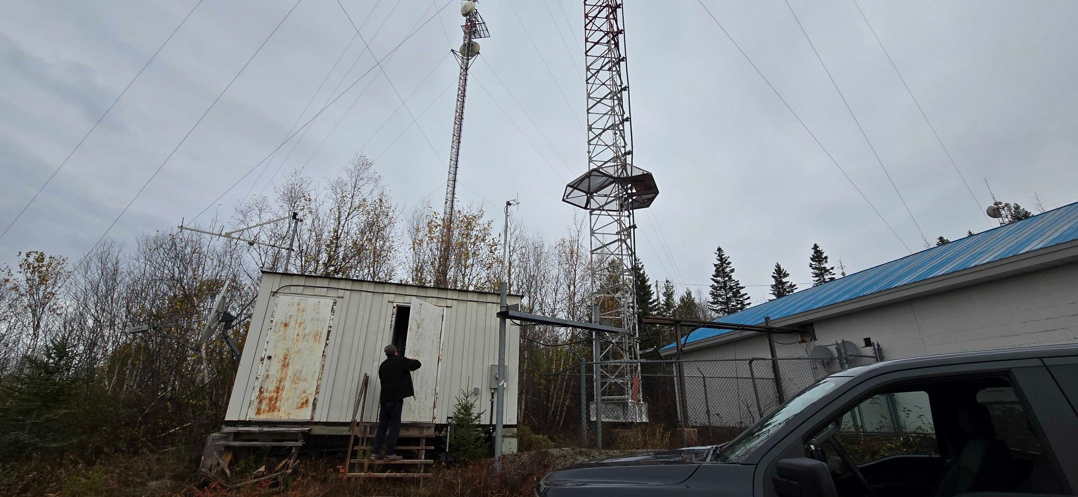 A guyed communications tower and equipment shelter at a bush site north of Wawa, Ontario, under an overcast October sky.